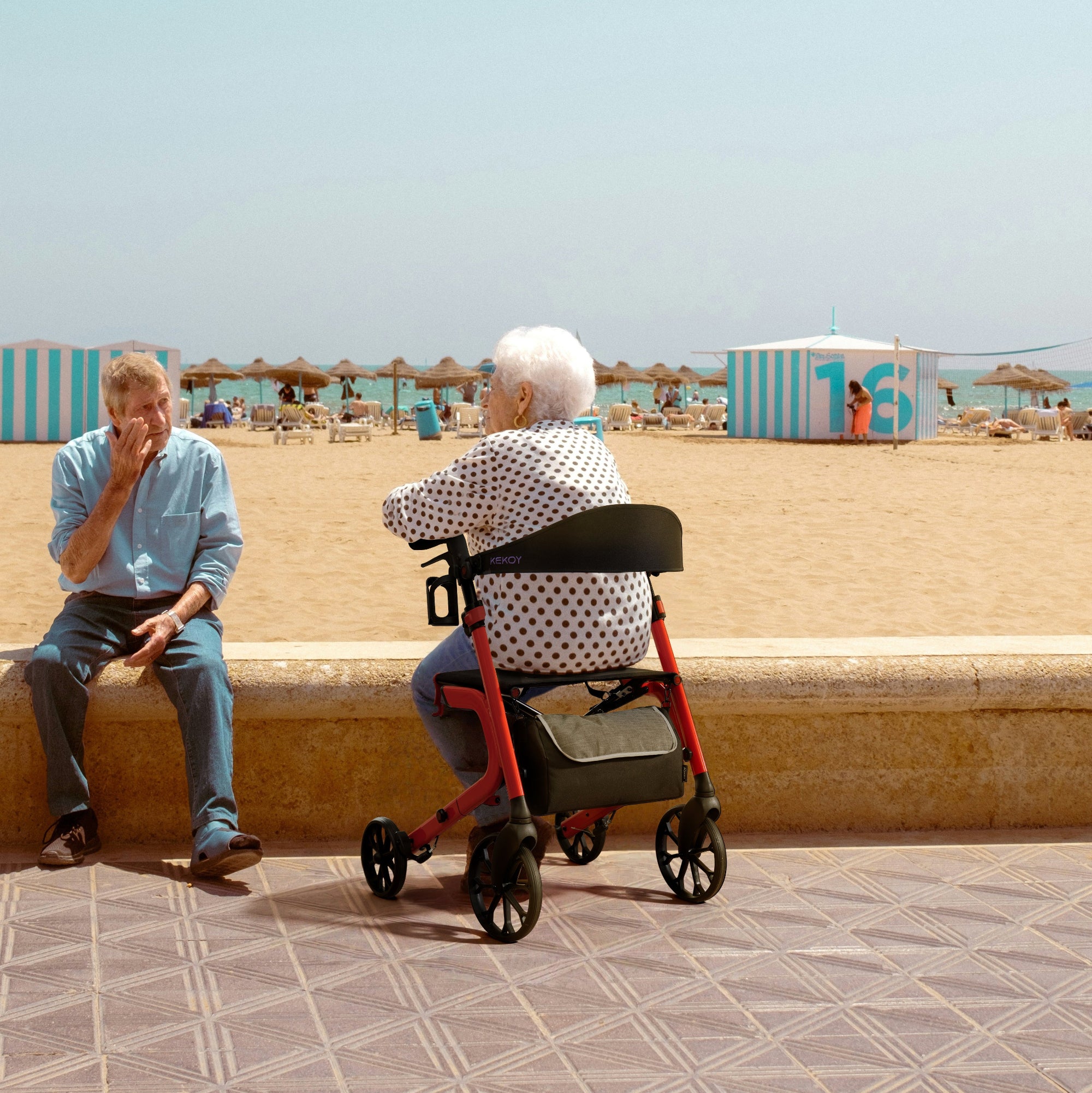 Two elderly people, one using a kekoy rollator sitting on a beachside pavement 