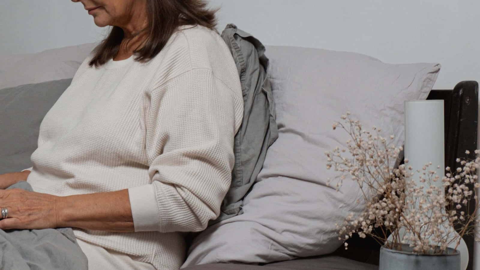 A woman sits on a bed with gray pillows and a gray throw blanket.